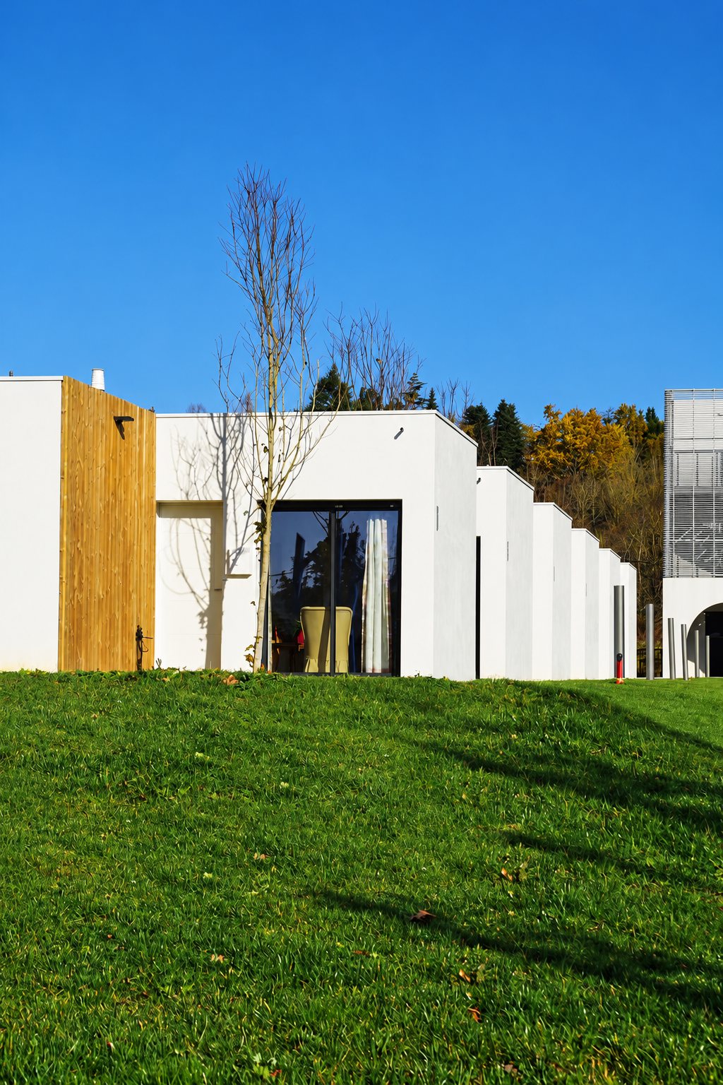 Façade d'un bâtiment moderne de plain-pied aux murs blancs et au panneau de bois, avec une grande baie vitrée, entouré d'une pelouse verte sous un ciel bleu.