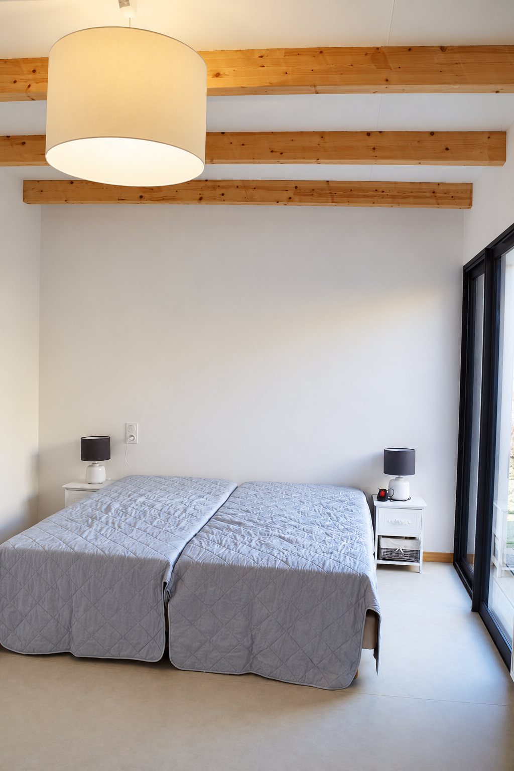 Chambre épurée avec un grand lit couvert d'un dessus-de-lit gris, deux tables de chevet, un lustre blanc et des poutres apparentes en bois.