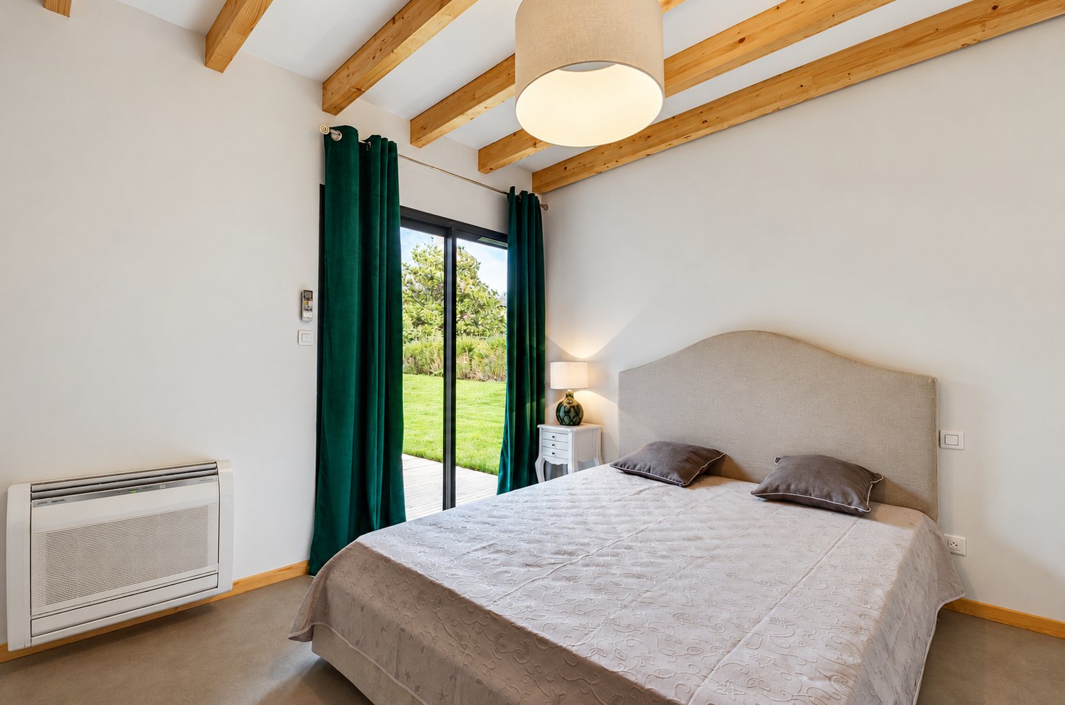 Chambre moderne avec un grand lit double, des rideaux verts devant une baie vitrée donnant sur un jardin, des poutres en bois au plafond et une climatisation murale.