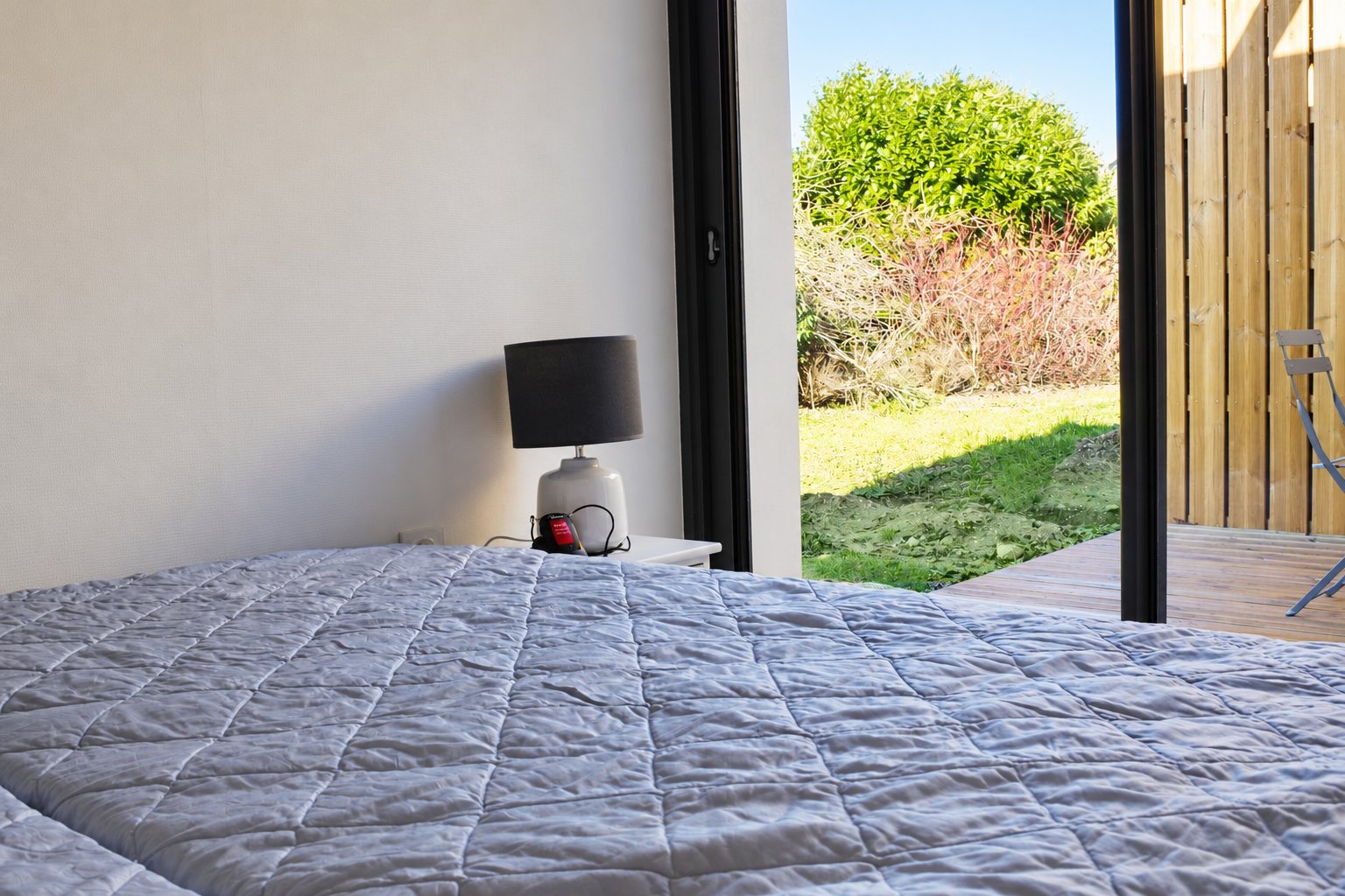 Une chambre lumineuse avec un lit couvert d'un couvre-lit matelassé gris, une lampe de chevet et une baie vitrée donnant sur un jardin et une terrasse en bois.