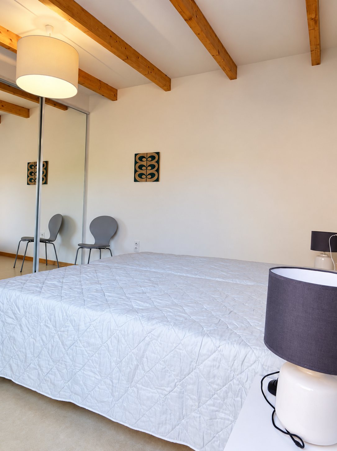 Chambre claire aux poutres en bois avec un grand lit couvert d'un dessus-de-lit blanc, des lampes de chevet grises et deux chaises grises reflétées dans une armoire à portes miroir.