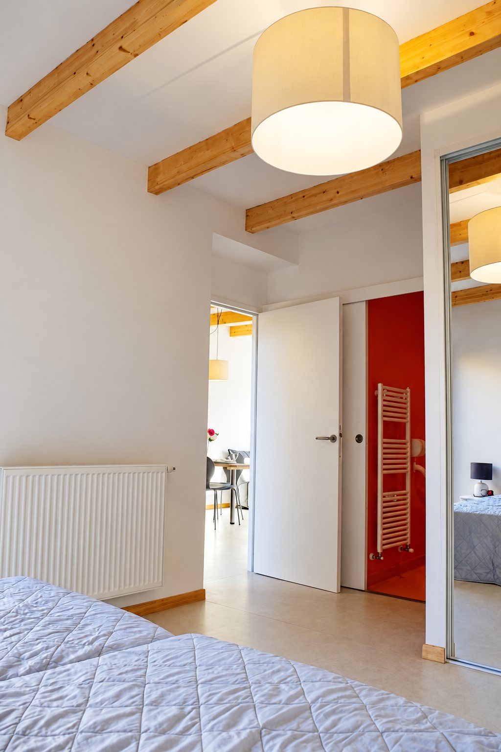 Chambre claire aux poutres en bois et à la grande suspension blanche, avec une porte ouverte sur une salle de bain rouge et un coin repas au fond.