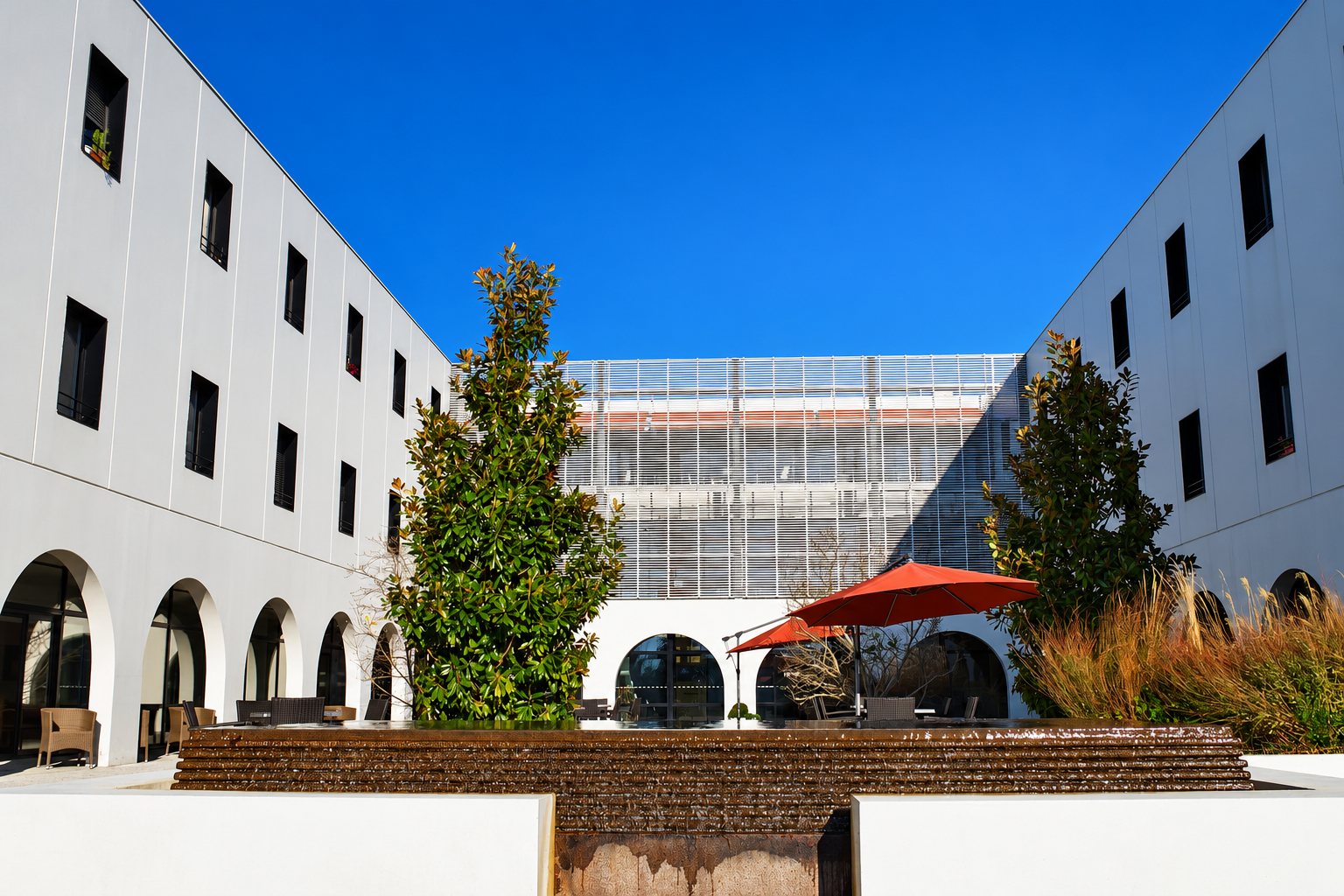 Une cour intérieure ensoleillée entourée de bâtiments blancs à arcades, avec une fontaine en cascade, des arbres et des parasols rouges sous un ciel bleu.
