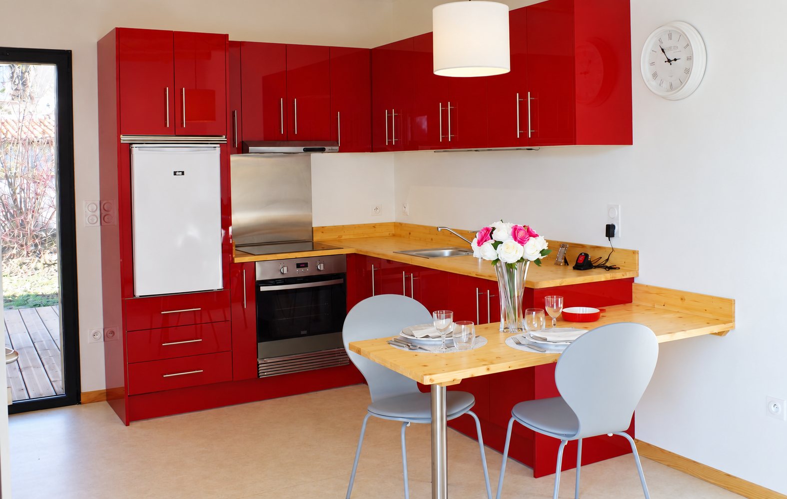 Cuisine moderne aux meubles rouge brillant et plan de travail en bois clair, avec une table dressée, deux chaises grises et un bouquet de fleurs.
