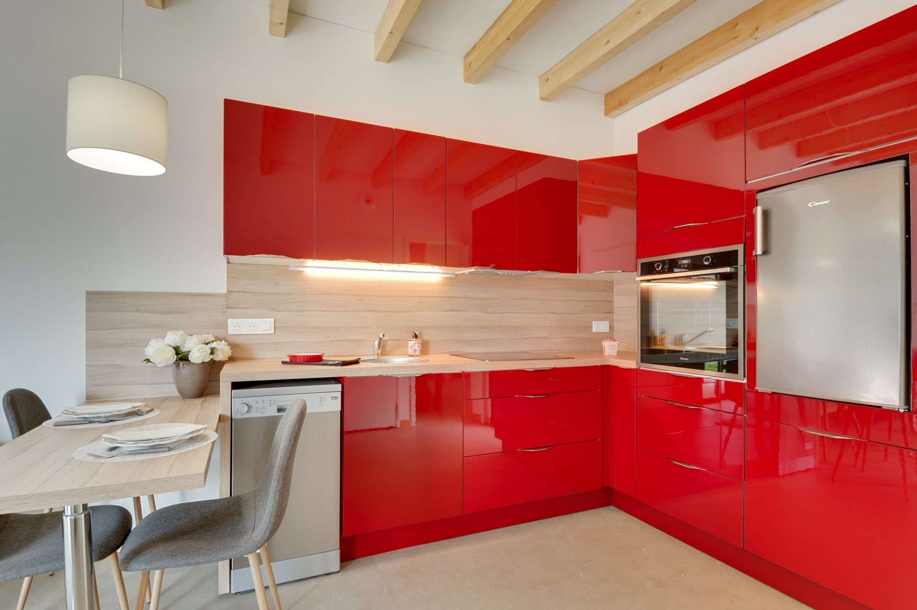 Cuisine équipée aux meubles rouge brillant, crédence en bois clair, électroménager inox et coin repas avec chaises grises sous des poutres apparentes.