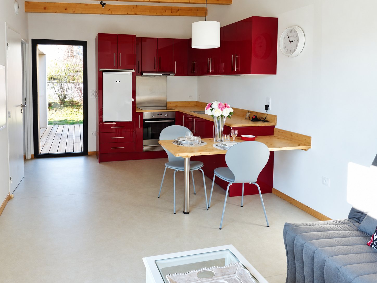 Intérieur lumineux d'un logement avec une cuisine rouge laquée, un plan de travail en bois formant table, deux chaises grises et une porte vitrée donnant sur une terrasse.