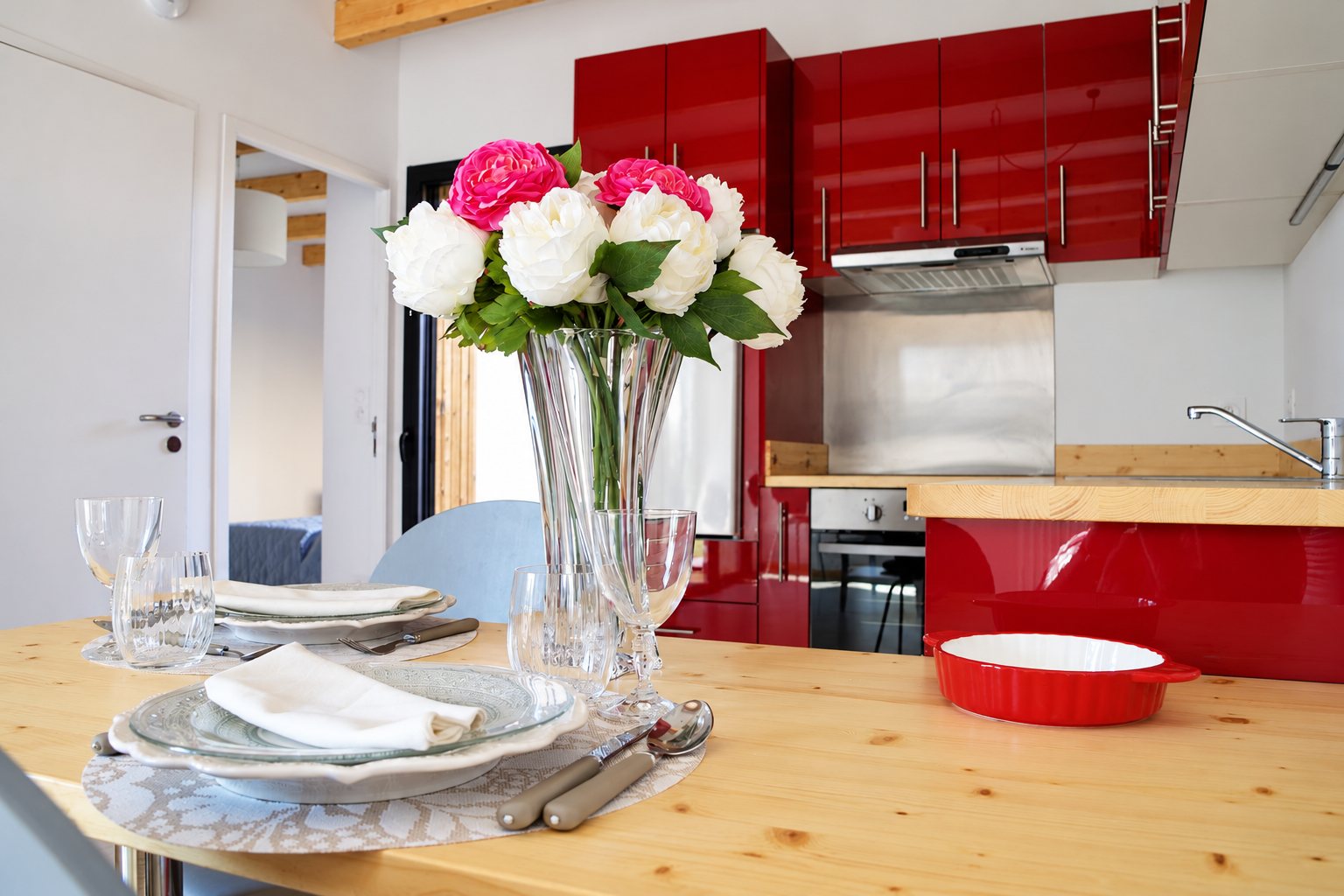 Une table en bois clair dressée avec assiettes, serviettes blanches et un vase de pivoines devant une cuisine ouverte aux meubles rouge laqué.
