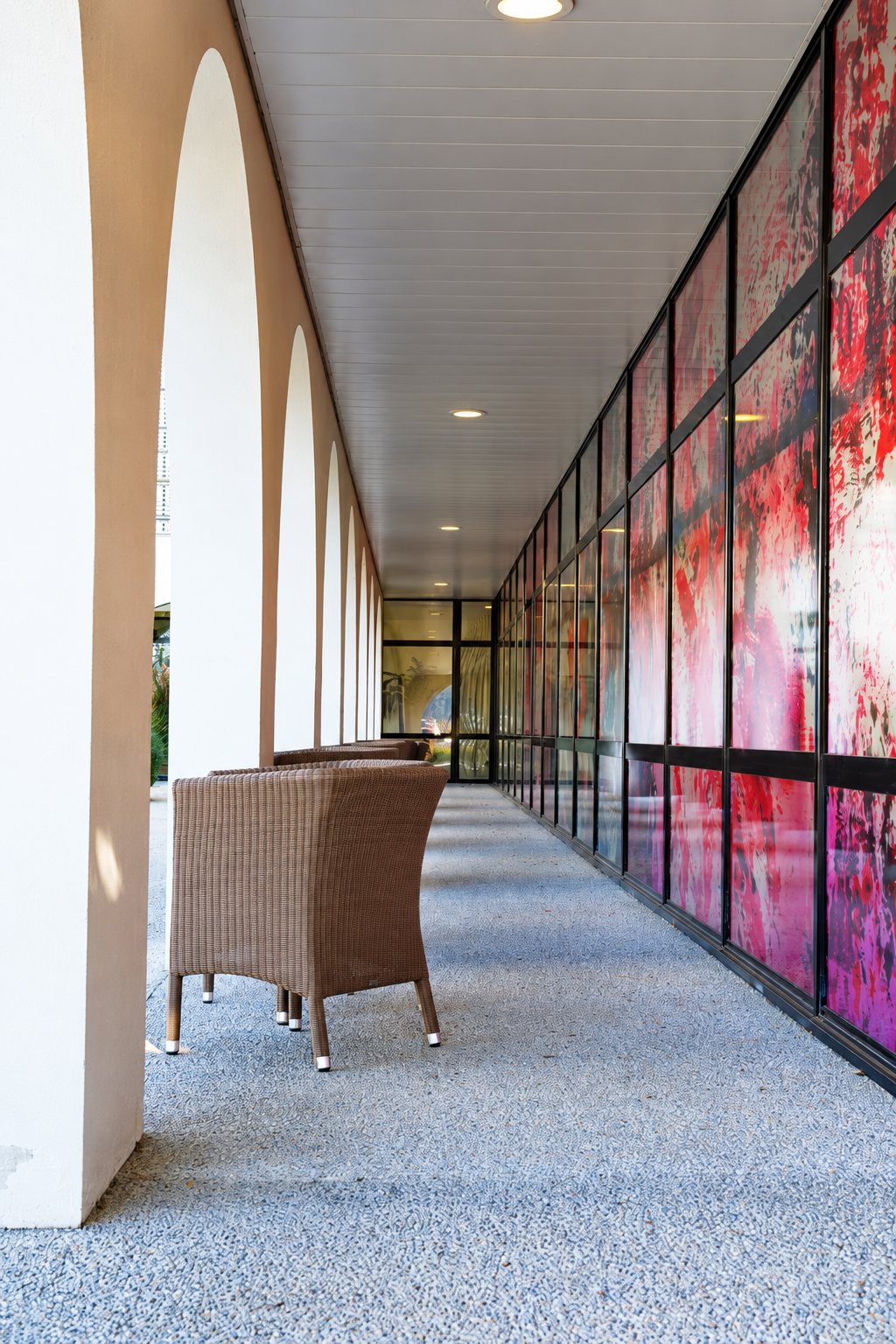Une longue galerie couverte à arcades blanches, où un fauteuil en rotin tressé fait face à une façade vitrée aux motifs rouges et roses.
