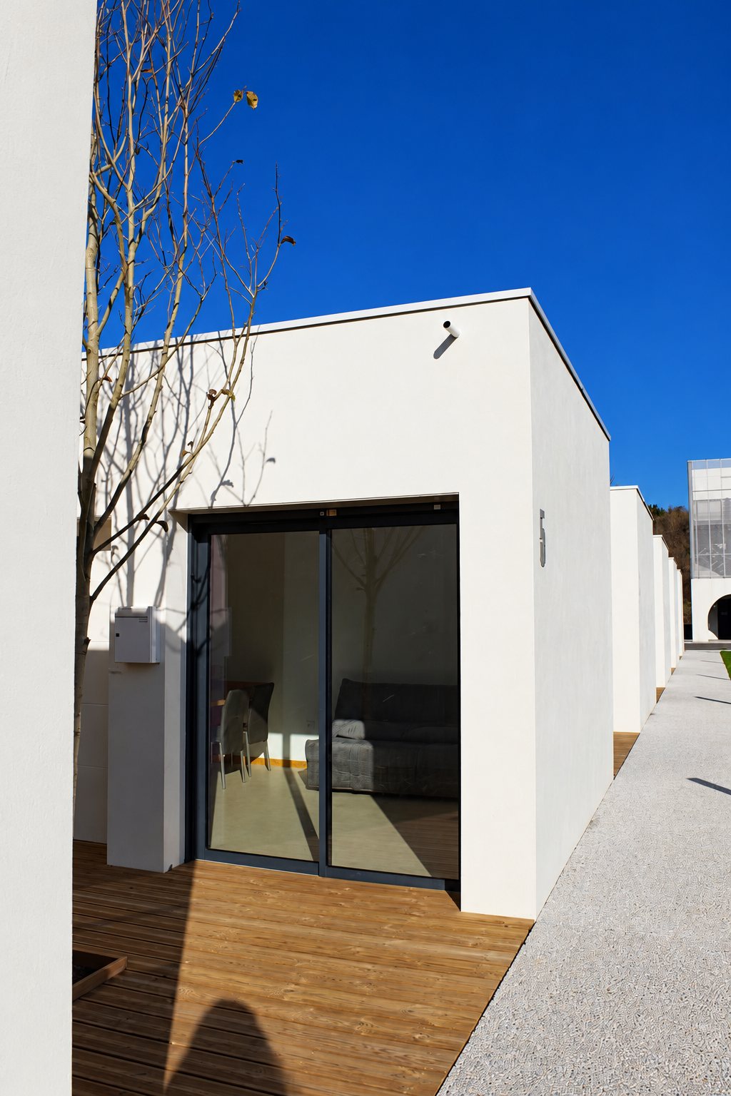 Façade d'un pavillon moderne aux murs blancs, avec une grande baie vitrée coulissante, une terrasse en bois et un arbre dénudé sous un ciel bleu.