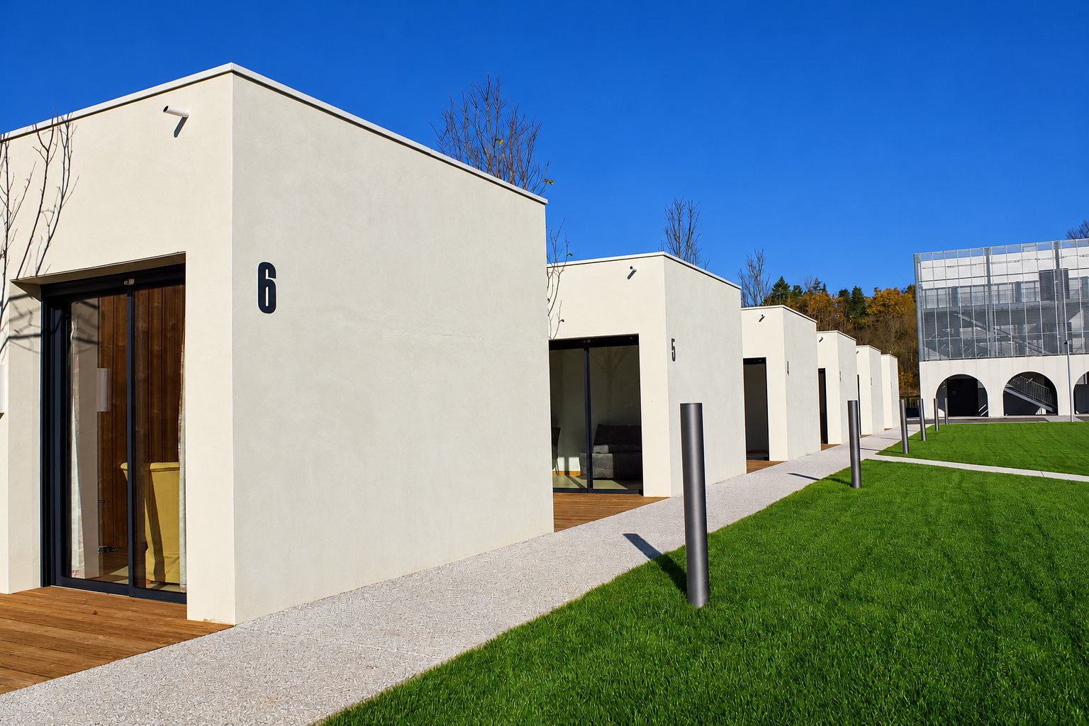Une rangée de petits pavillons modernes aux murs blancs, numérotés, avec terrasses en bois, allée en gravier clair et pelouse verte sous un ciel bleu.