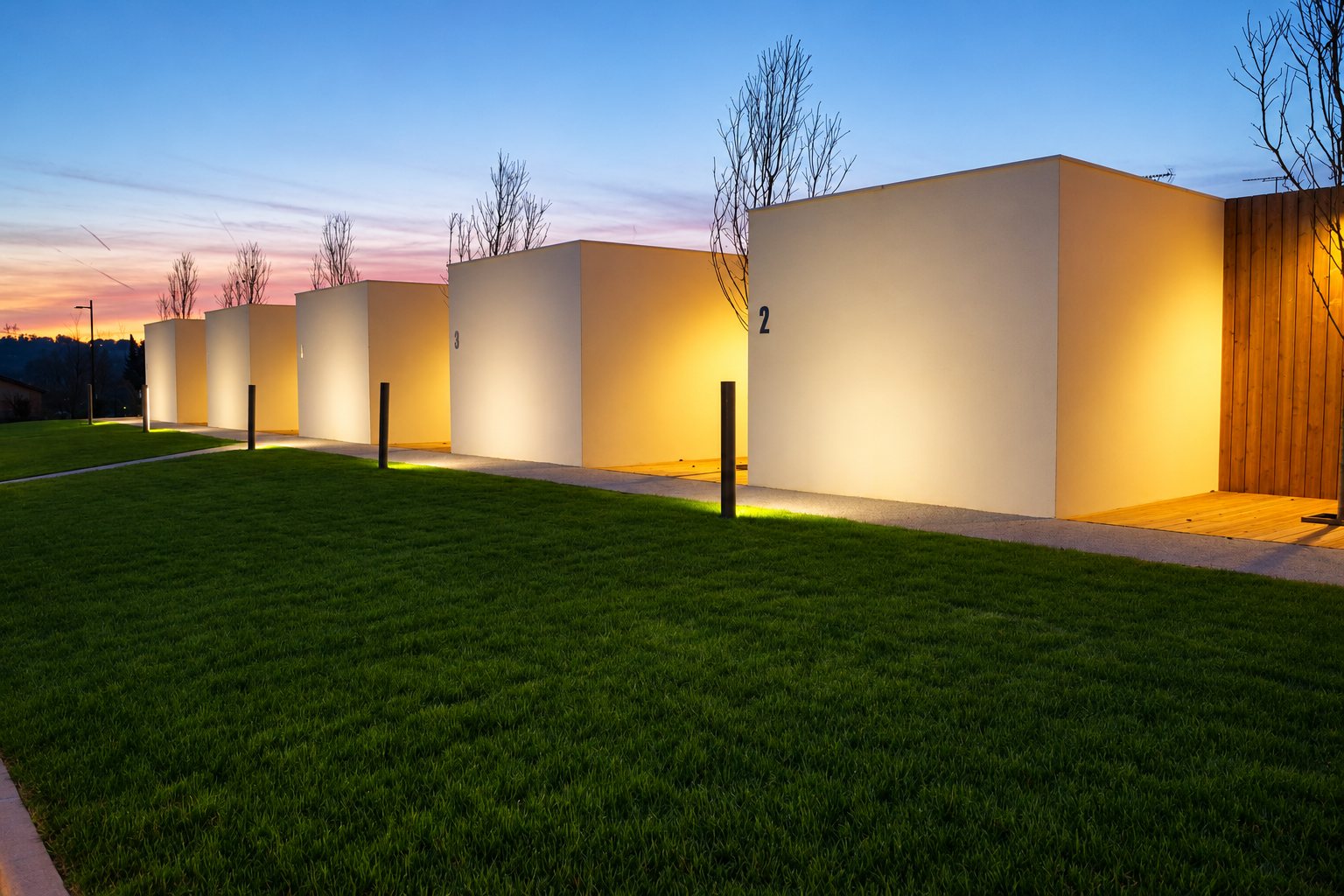 Au crépuscule, une rangée de pavillons cubiques blancs aux façades éclairées en lumière chaude borde une pelouse, le numéro 2 visible sur le premier.