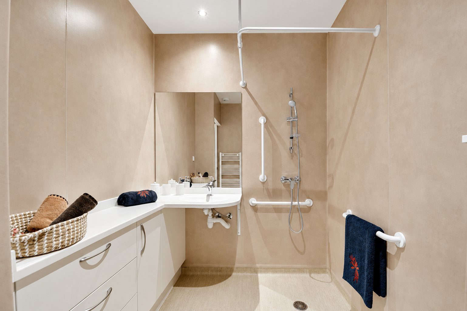 Salle de bain aux murs beige rosé avec douche à l'italienne, barres d'appui blanches, lavabo suspendu et meuble blanc avec serviettes.