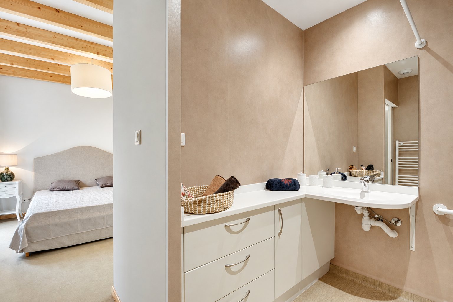 Salle de bain aux murs beige rosé avec meuble blanc, lavabo suspendu et barre d'appui, donnant sur une chambre avec lit et poutres apparentes.