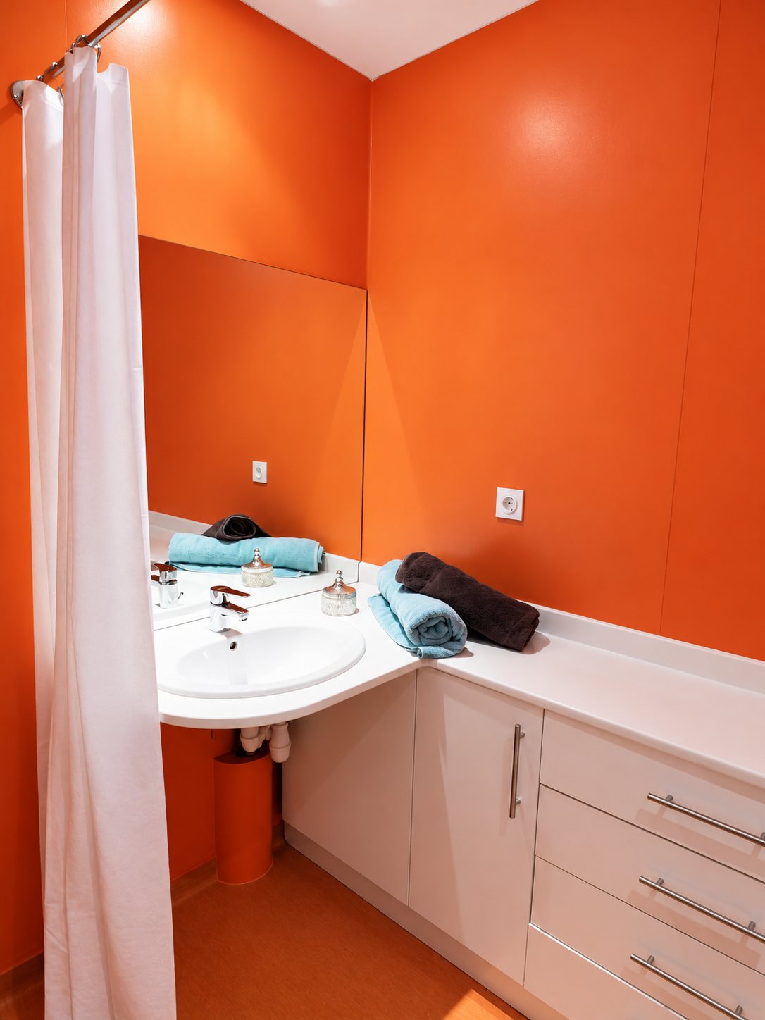 Salle de bain aux murs orange avec un plan vasque blanc, un rideau de douche blanc et des serviettes bleues et brunes soigneusement roulées.