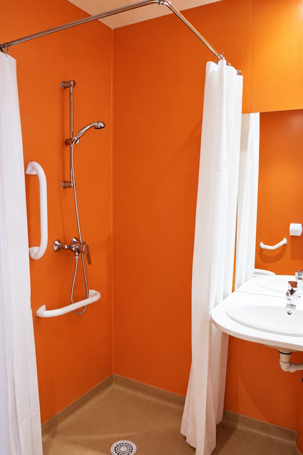 Salle de bain aux murs orange avec une douche équipée de barres d'appui blanches, de rideaux blancs et d'un lavabo suspendu avec miroir.