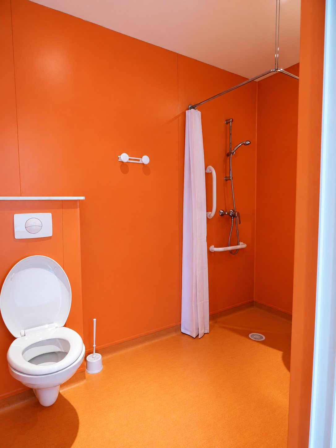 Salle de bains aux murs et au sol orange avec WC suspendu, douche de plain-pied, rideau blanc et barres d'appui.