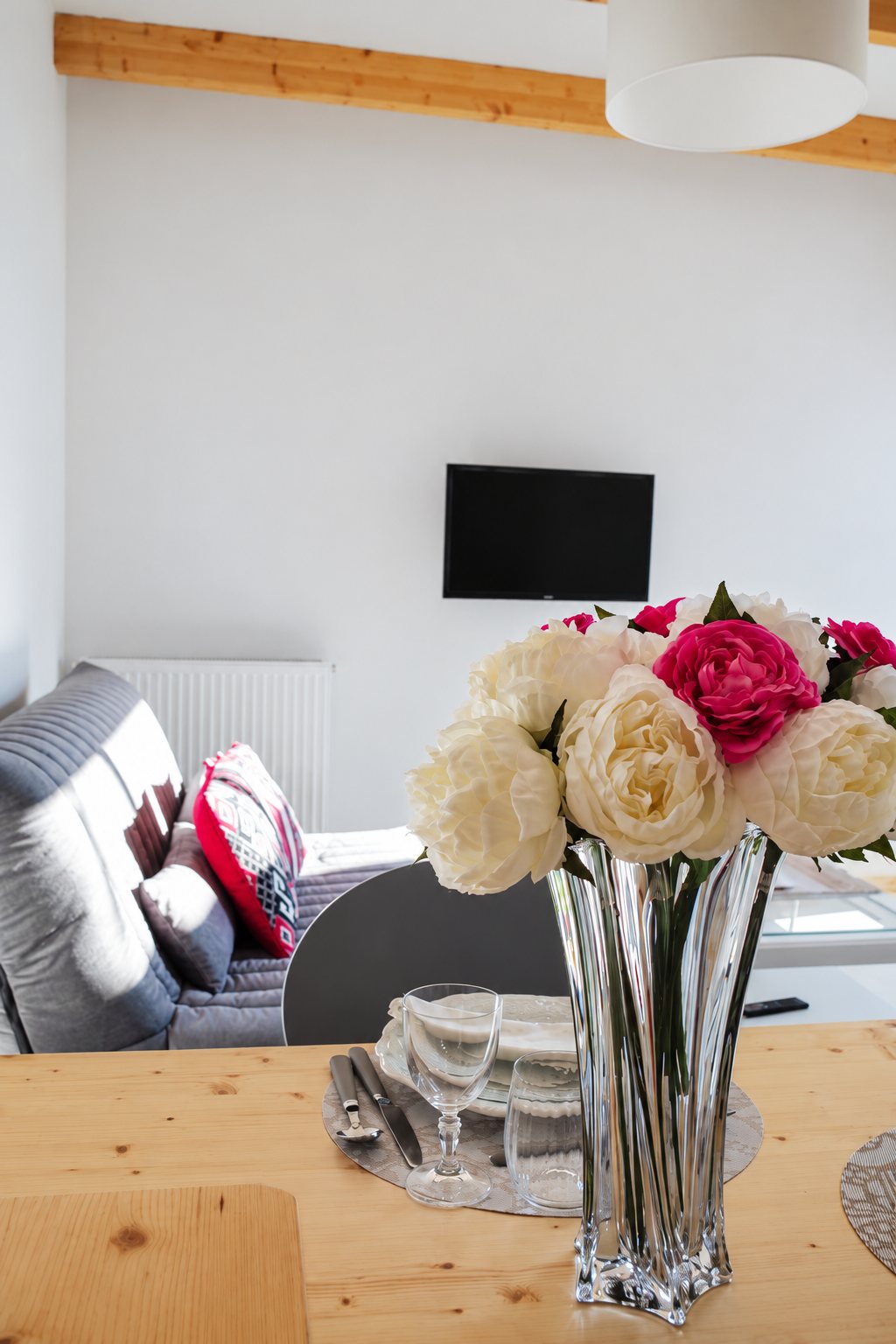 Un vase en cristal rempli de pivoines blanches et rouges est posé sur une table en bois dressée, devant un canapé gris et un téléviseur mural dans un salon clair.