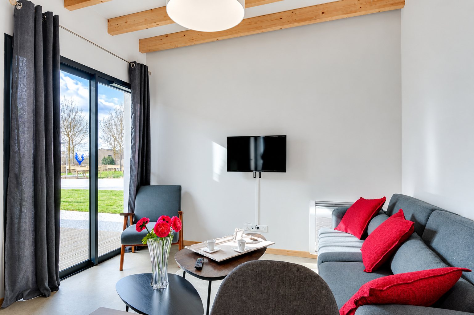 Un salon moderne aux murs blancs avec un canapé gris orné de coussins rouges, une table basse, un bouquet de fleurs roses et une grande baie vitrée donnant sur un jardin.