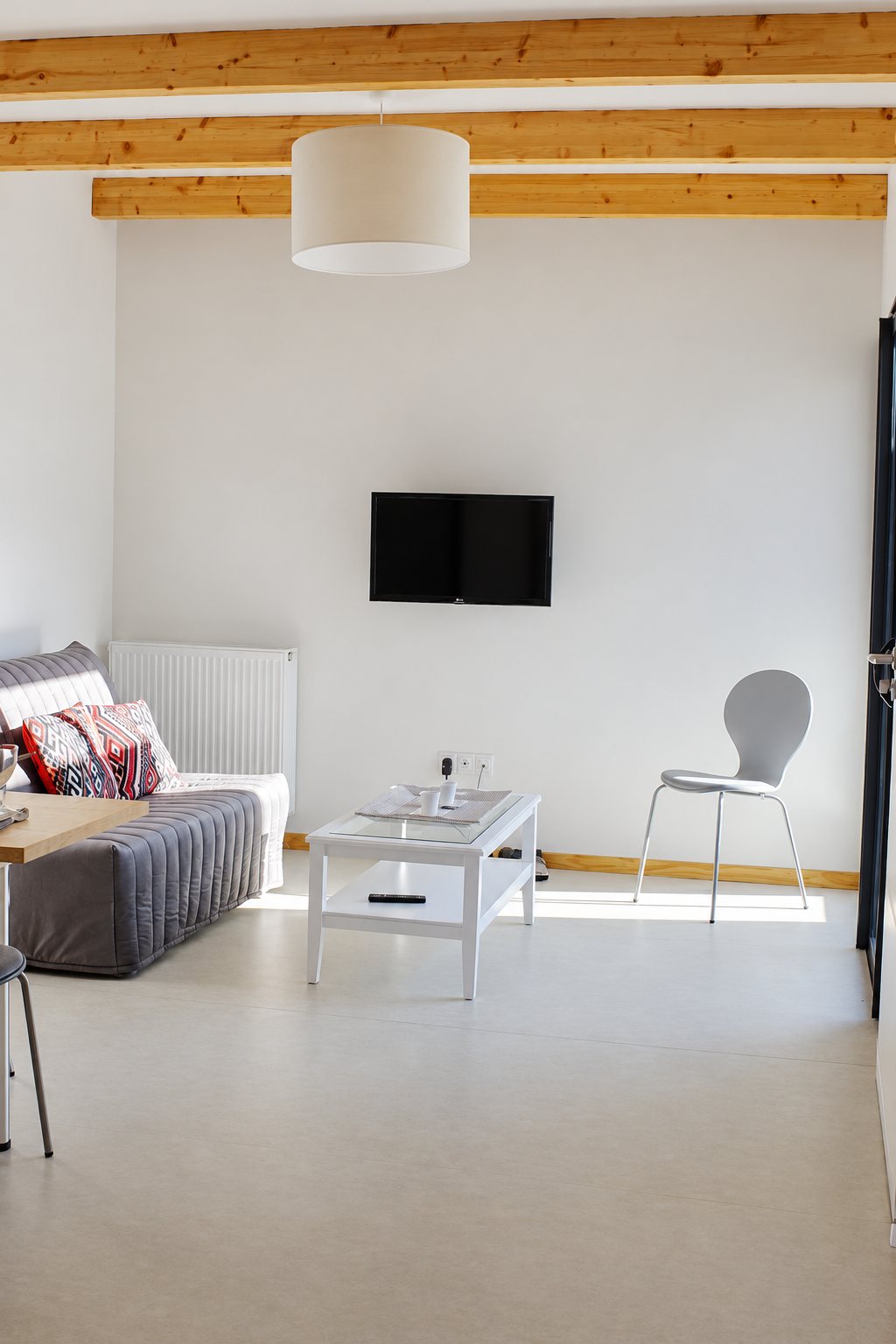Salon moderne et lumineux avec un canapé gris, des coussins colorés, une table basse blanche, une chaise grise, une télévision murale et des poutres apparentes en bois.