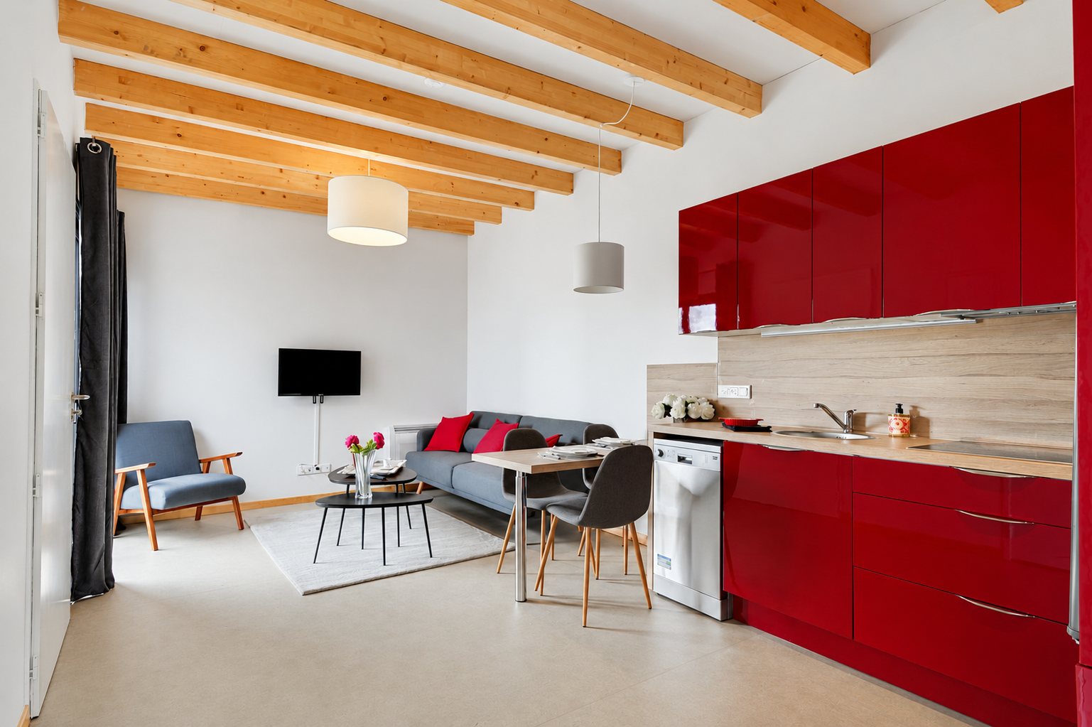Pièce de vie moderne avec cuisine équipée rouge laquée, canapé gris, fauteuil vintage, table à manger et plafond à poutres en bois clair.