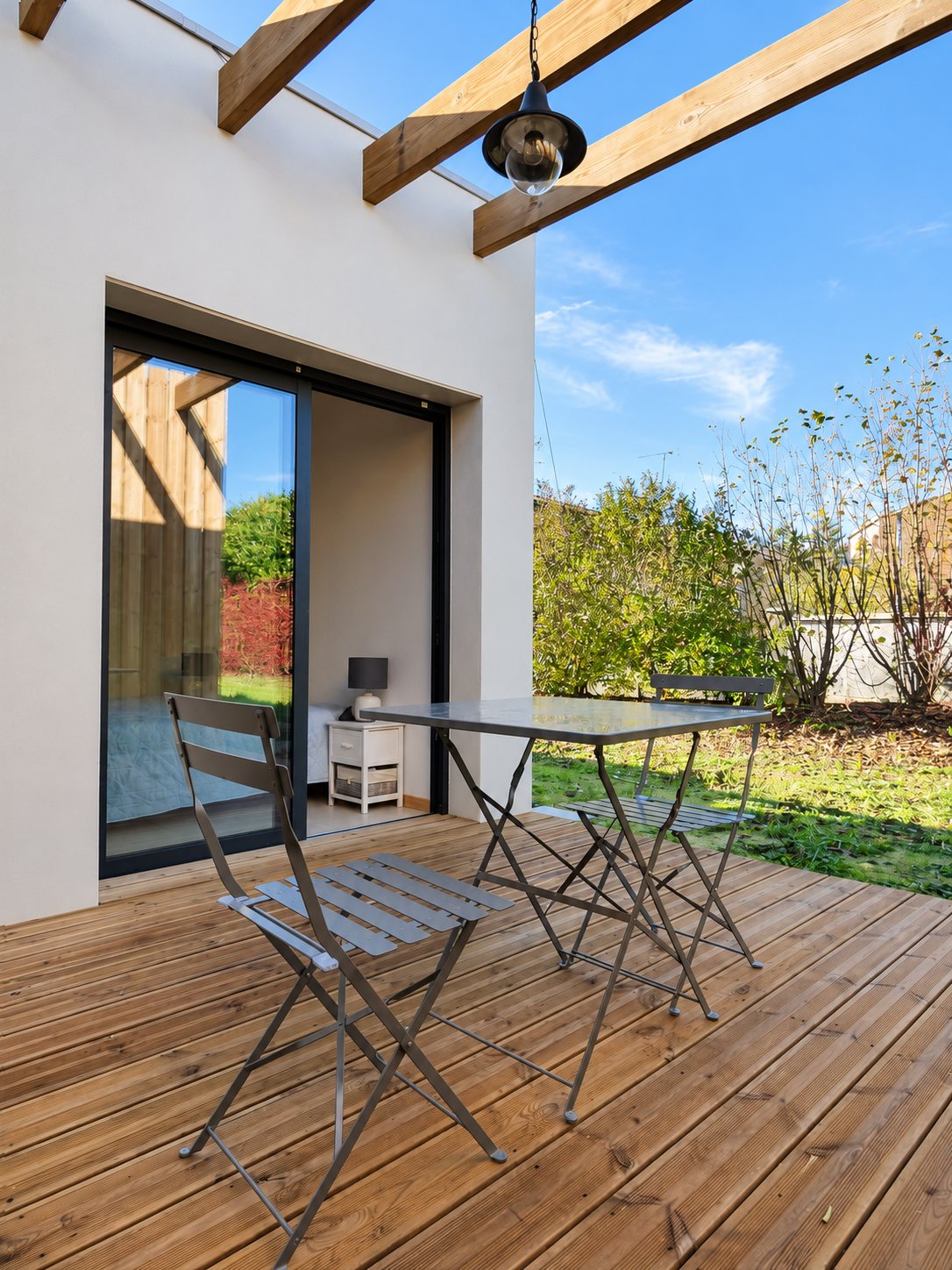 Une terrasse en bois avec pergola, une table et deux chaises pliantes grises devant une baie vitrée coulissante donnant sur une chambre, avec un jardin en arrière-plan.